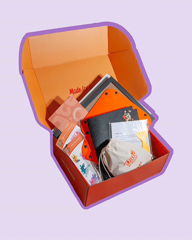 Angled view of Date in a Crate Puzzle Crate on a lavender background with a purple outline, featuring a puzzle guide, themed puzzle date stickers, felt puzzle mat, two orange puzzle piece holders, glue sheets with a plastic scraper and a canvas bag containing a custom puzzle with the Date in a Crate logo.