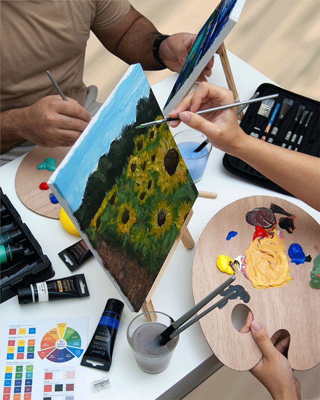 Close-up of two people painting on canvases set on small wooden easels, one featuring a sunflower field. A wooden palette with mixed acrylic paints, paintbrushes in water, paint tubes, a colour wheel, and other painting supplies are on the table.
