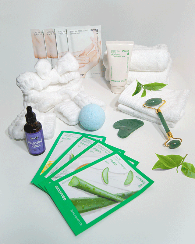 Flat lay of spa products including Innisfree sheet masks, Green Tea cleansing foam, Relaxation Ritual massage oil, jade roller and gua sha, white towels, fluffy headband, blue bath bomb, and green leaves on a white background.