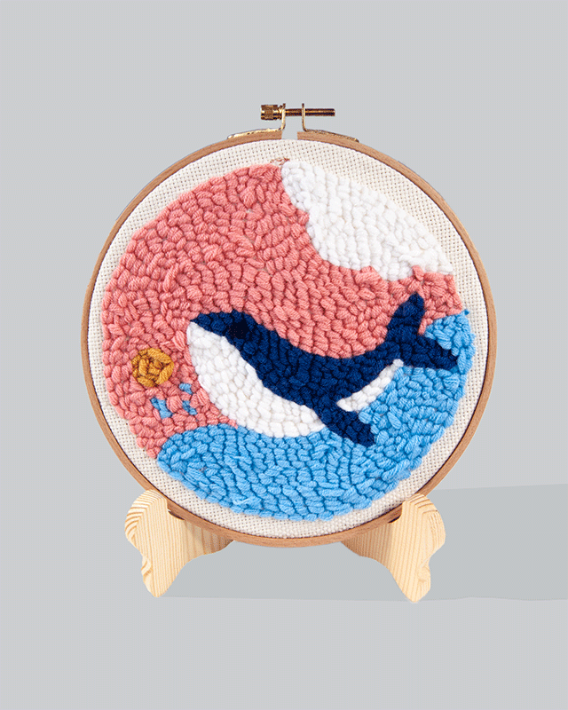 Embroidery hoop featuring a dark blue whale swimming across a blue ocean wave with a pink background, and a small golden sun in the corner.