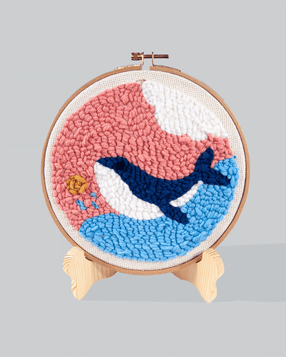 Embroidery hoop featuring a dark blue whale swimming across a blue ocean wave with a pink background, and a small golden sun in the corner.
