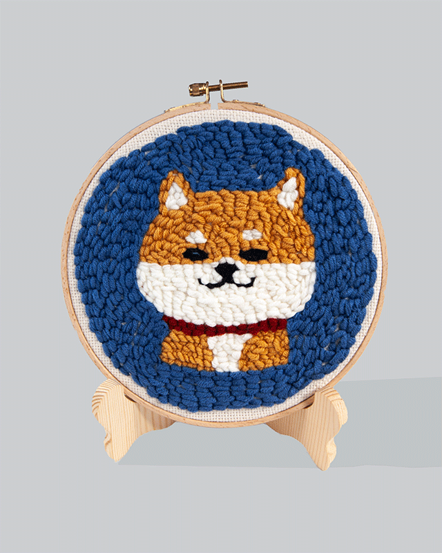 Embroidery hoop featuring a cute Shiba Inu dog with orange fur, white face and chest, and a red collar, set against a blue stitched background.