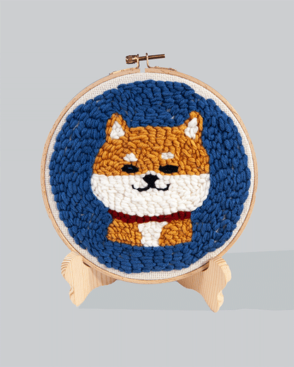 Embroidery hoop featuring a cute Shiba Inu dog with orange fur, white face and chest, and a red collar, set against a blue stitched background.