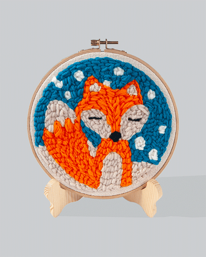 Embroidery hoop featuring an orange fox with eyes closed on a blue stitched background with white snow-like details.