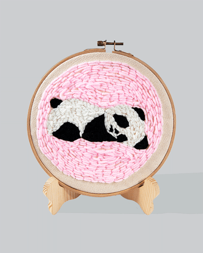 Embroidery hoop with a sleepy panda design on a pink stitched background.