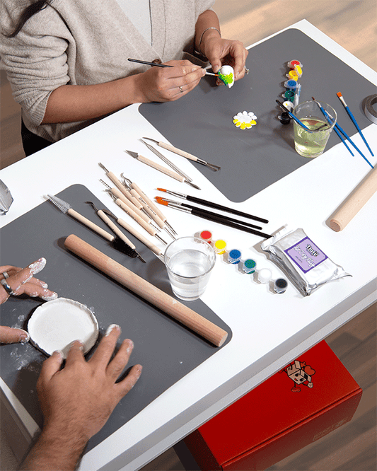 Couple working with white air-dry clay and painting small clay creations on grey mats, surrounded by sculpting tools, paintbrushes, rolling pin, and colourful paint pots.