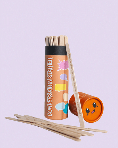 Conversation Starter game tube with orange lid open, showing wooden sticks inside with printed questions, one leaning against the tube and several spread on the surface, set on a light purple background.