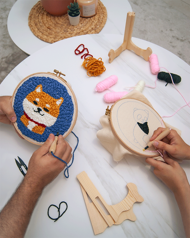 Two people working on embroidery hoops, one stitching a Shiba Inu design and the other starting a panda pattern, with yarn, tools, and wooden stands on a white table.