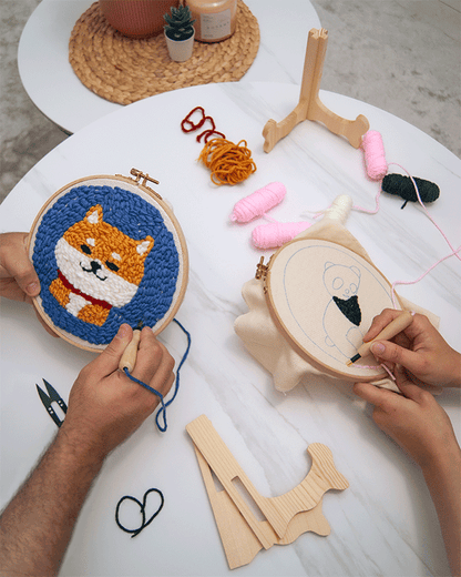 Two people working on embroidery hoops, one stitching a Shiba Inu design and the other starting a panda pattern, with yarn, tools, and wooden stands on a white table.