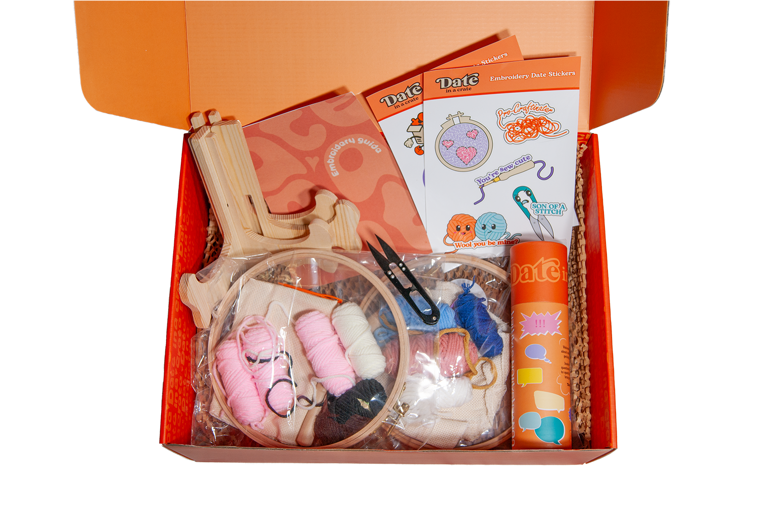 Open Date in a Crate Embroidery Crate box containing embroidery hoops with fabric and pre-sorted threads, wooden display stands, small black snips, embroidery guide booklet, themed stickers, and a conversation starter tube.