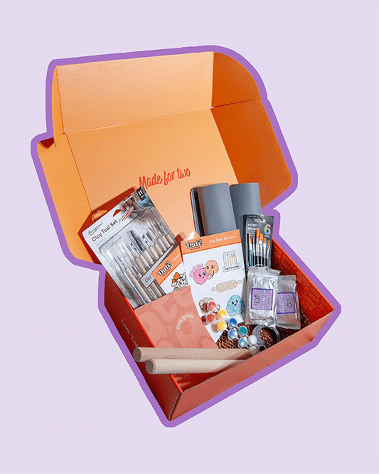 Angled view of Date in a Crate Clay Crate on a lavender background with a purple outline, showing clay tool set, grey silicone mats, paintbrushes, paint pots, wooden rolling pins, clay guide, clay-themed stickers, two packs of 250g air dry clay, and magnet tape.