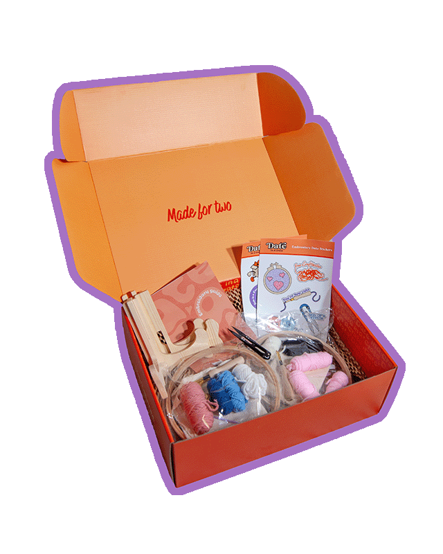 Angled view of Date in a Crate Embroidery Crate on a lavender background with a purple outline, showing wooden display stands, embroidery hoops with pre-drawn designs, colourful yarn, embroidery needles, scissors, an embroidery guide, and themed stickers