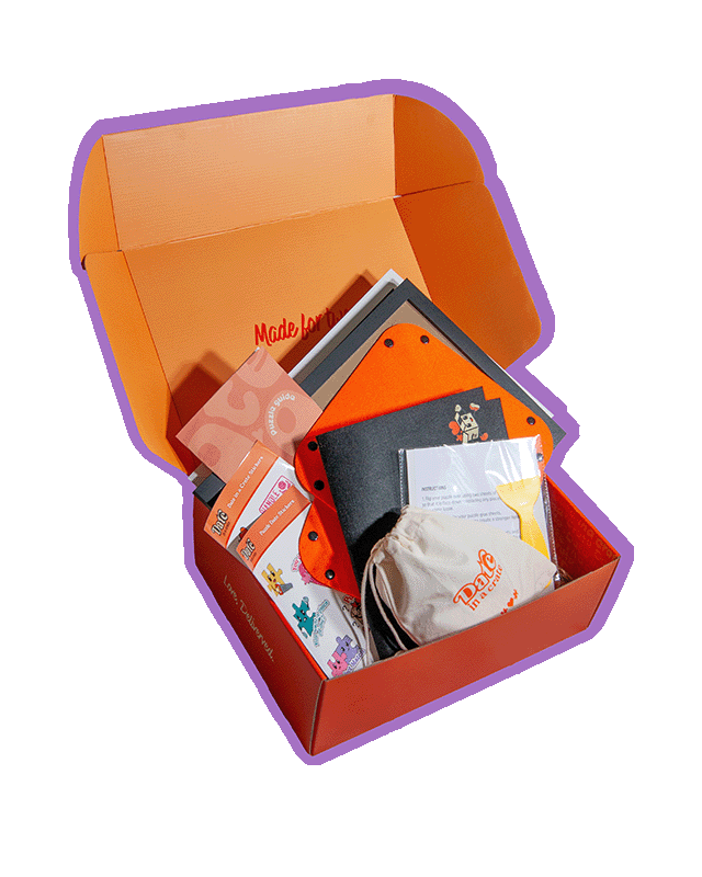 Angled view of Date in a Crate Puzzle Crate on a lavender background with a purple outline, featuring a puzzle guide, themed puzzle date stickers, felt puzzle mat, two orange puzzle piece holders, glue sheets with a plastic scraper and a canvas bag containing a custom puzzle with the Date in a Crate logo.