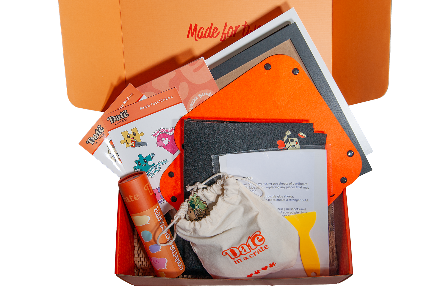 Open Date in a Crate Puzzle Crate box showing contents including puzzle stickers, instruction sheets, orange and black cardboard pieces, conversation starter tube, small drawstring bag, and yellow puzzle glue applicator.