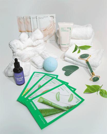 Flat lay of spa products including Innisfree sheet masks, Green Tea cleansing foam, Relaxation Ritual massage oil, jade roller and gua sha, white towels, fluffy headband, blue bath bomb, and green leaves on a white background.